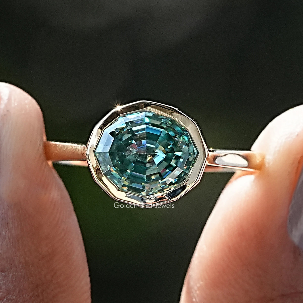 Front view of Blue Green Moissanite Step Cut Oval Solitaire Ring in two fingers