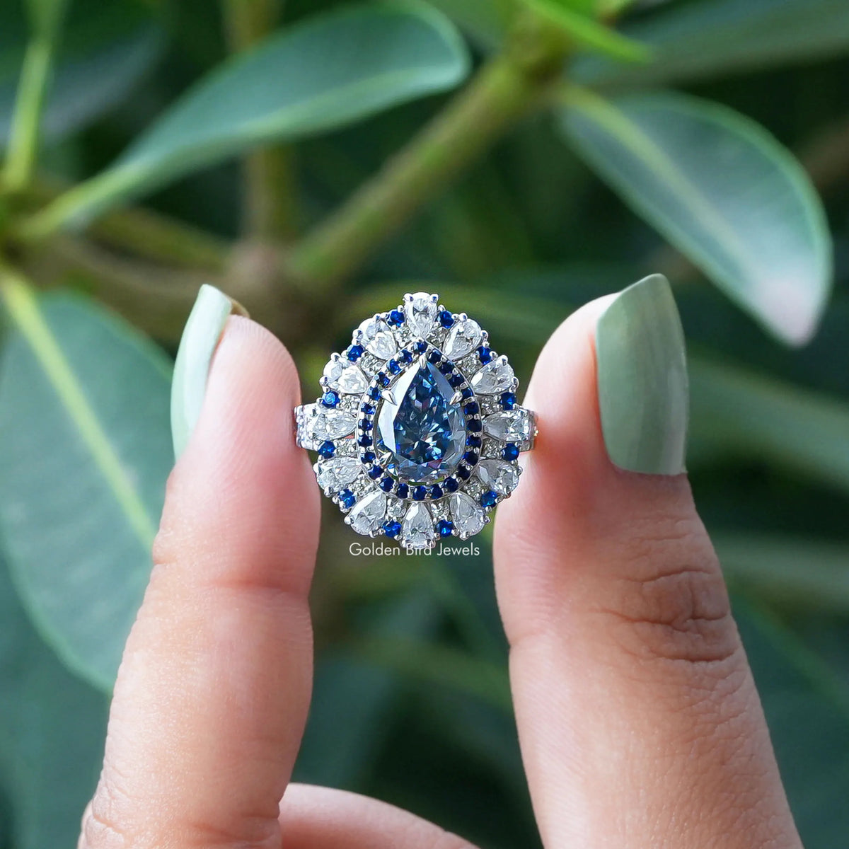 Two fingers holding the Blue Pear Cut Moissanite Halo Engagement Ring