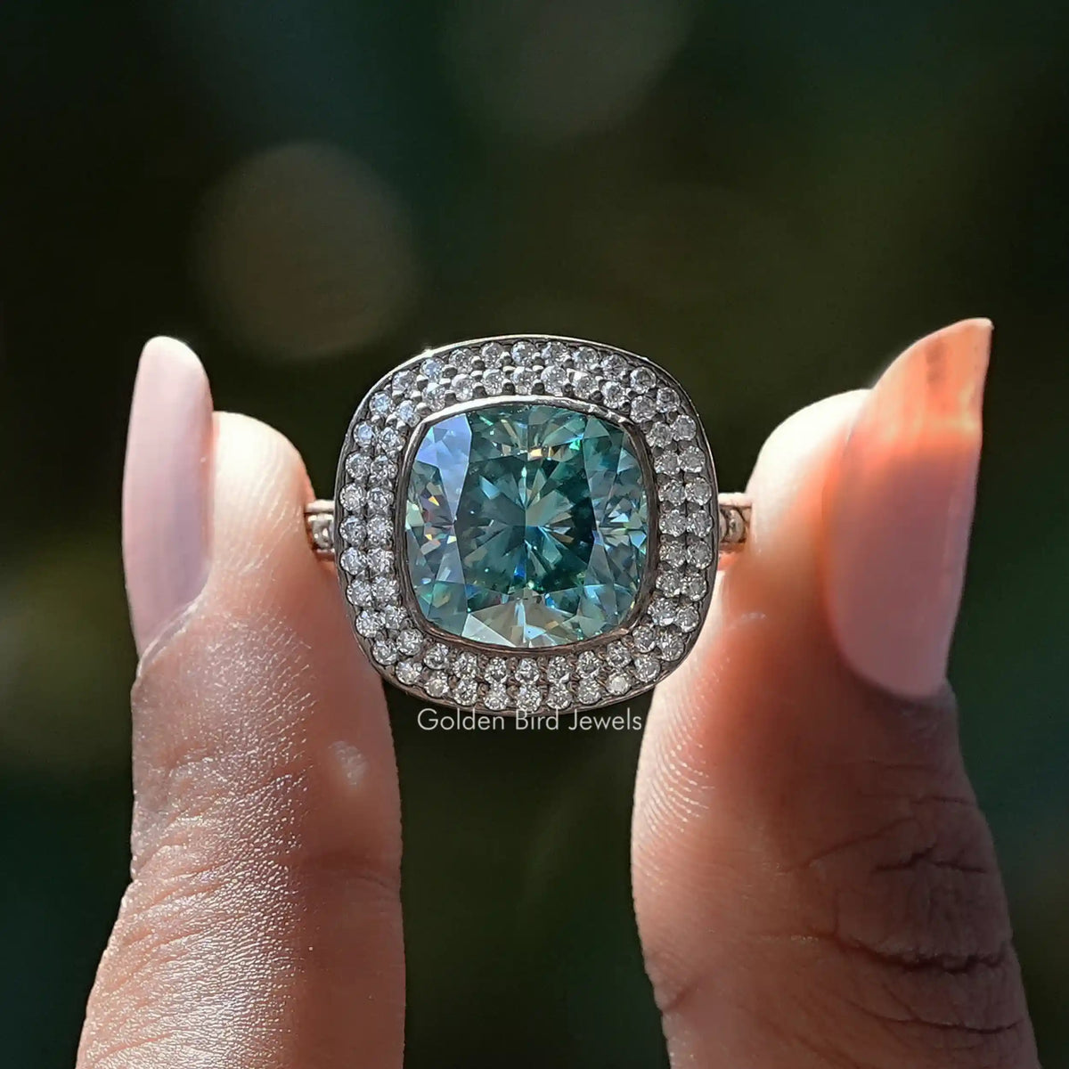 Two fingers holding the Blue Green Cushion Cut Moissanite Double Halo Ring