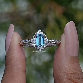 Two fingers holding the Emerald Cut Halo Engagement Ring In white Gold