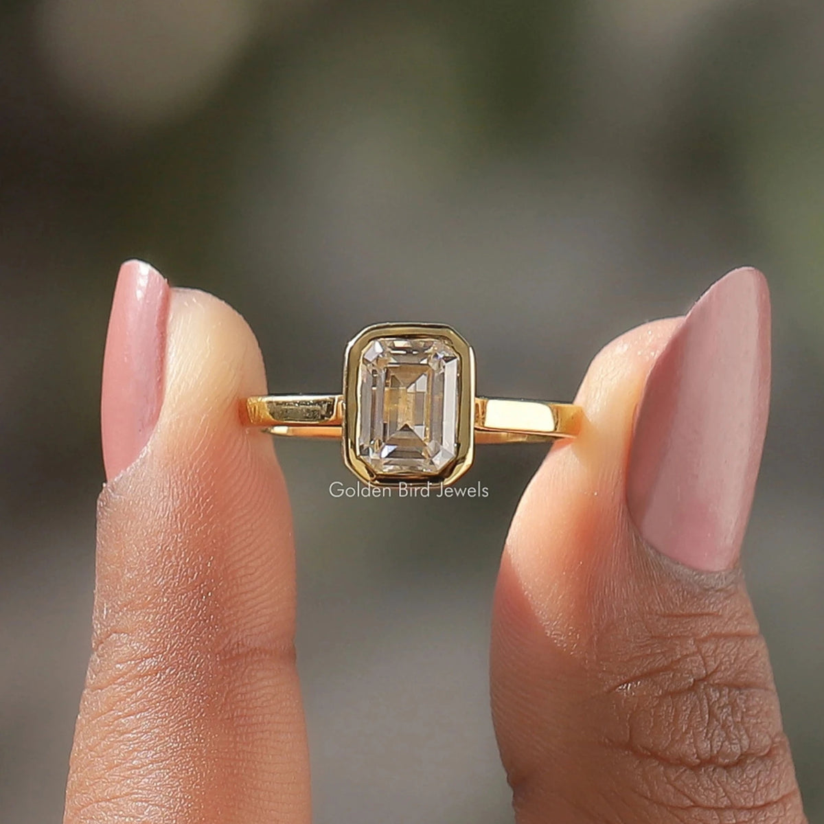 Two fingers holding the Emerald Cut Moissanite Bezel Set Ring