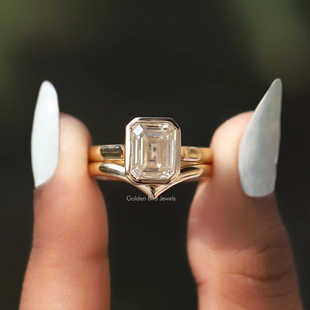 [In two finger front view of emerald cut moissanite solitaire bezel set ring]-[Golden Bird Jewels]