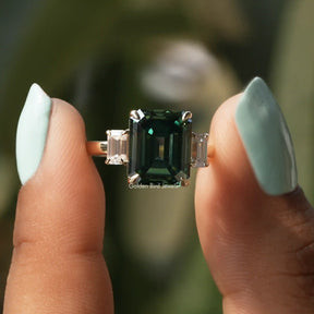 Two fingers holding the Green Emerald Cut Three Stone Engagement Ring