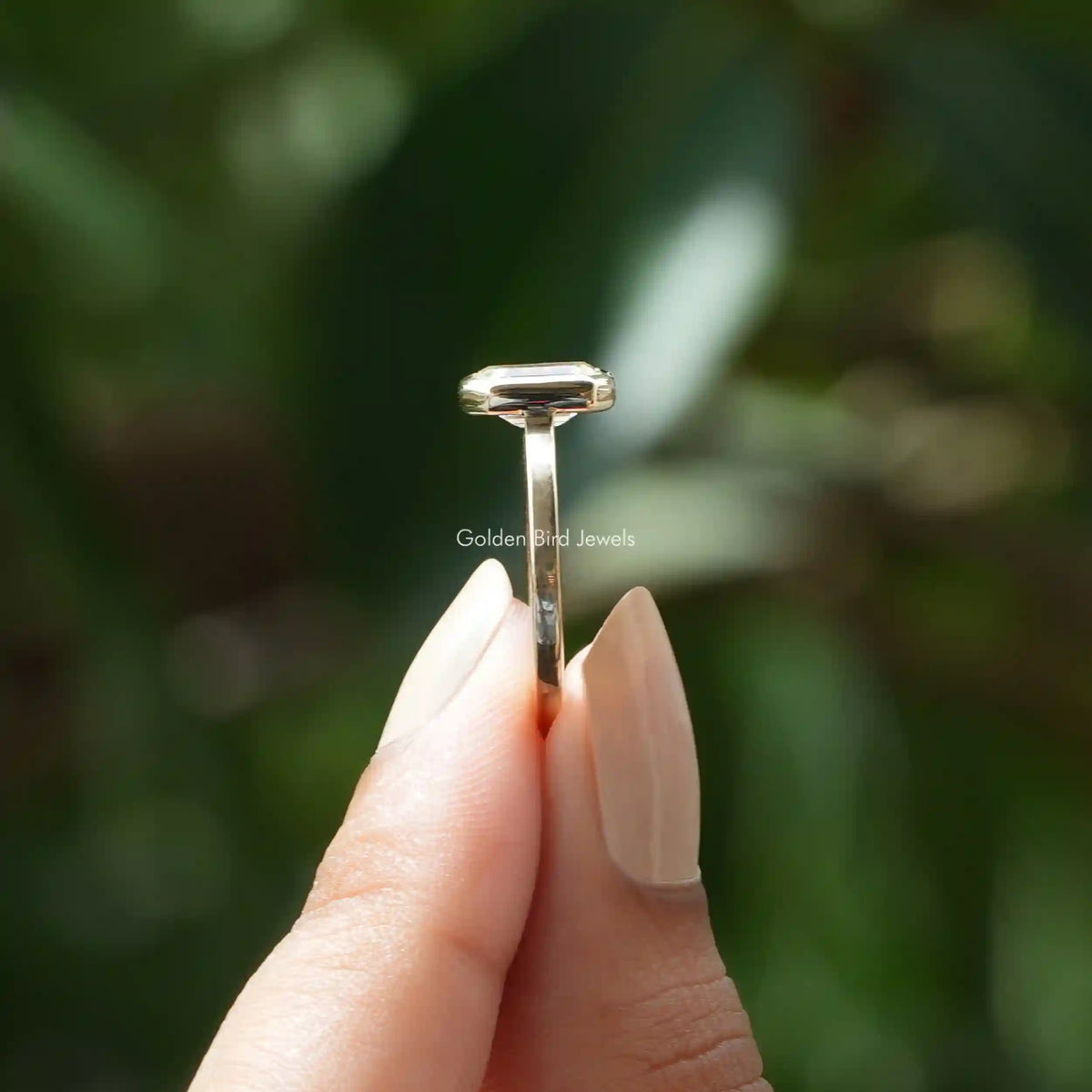 Side view of emerald cut bezel set ring elegantly held with two fingers.