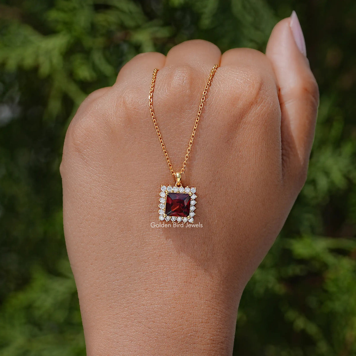 Ruby-red square pendant with clear halo stones on gold chain necklace, worn on hand