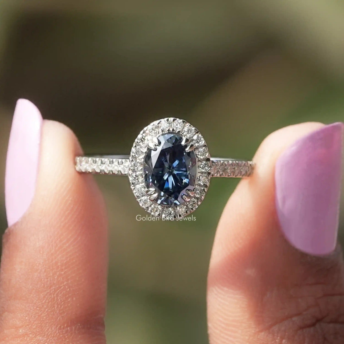 Two fingers holding the Sapphire Blue Oval Cut Halo Engagement Ring